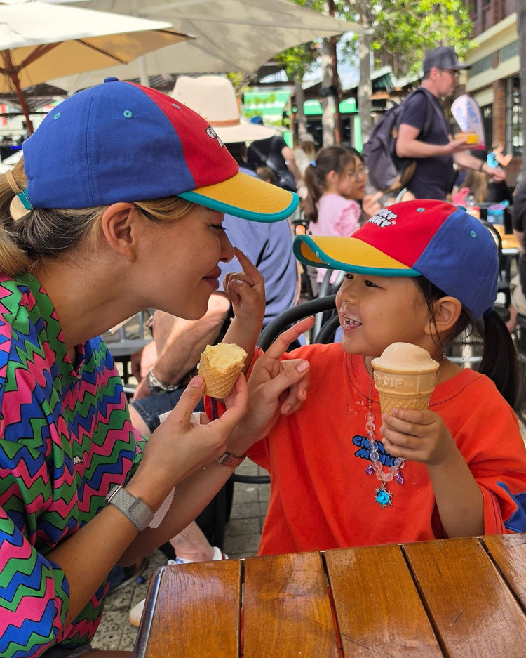 A woman and a young girl sharing an ice cream cone at an outdoor cafe table, both wearing matching red, blue, and yellow baseball caps.