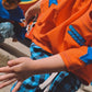 A child wearing a bright orange t-shirt with blue star patches and blue plaid pants, dancing in front of a colorful graffiti wall. Another child is visible in the background.