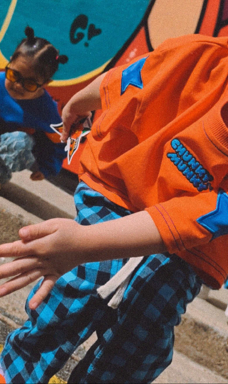 A child wearing a bright orange t-shirt with blue star patches and blue plaid pants, dancing in front of a colorful graffiti wall. Another child is visible in the background.
